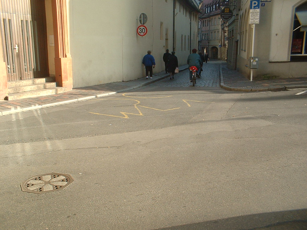 A yellow warchalking open-node symbol marked on the tarmac of a street corner in a European town, with pedestrians and a cyclist in the background